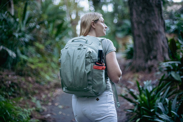 A woman walking a leafy path wearing the emmèton Metro 20L anti-theft backpack in sage green, an umbrella in the side pocket