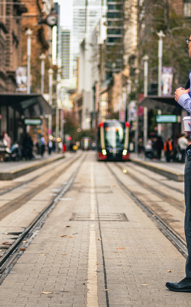 Man with Urban 17.5L backpack, walking on a city street with tram tracks and urban buildings in the background.