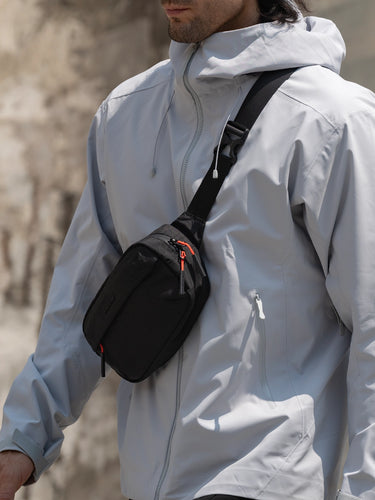 Person wearing a light gray jacket with a black sling bag over one shoulder against a blurred background.